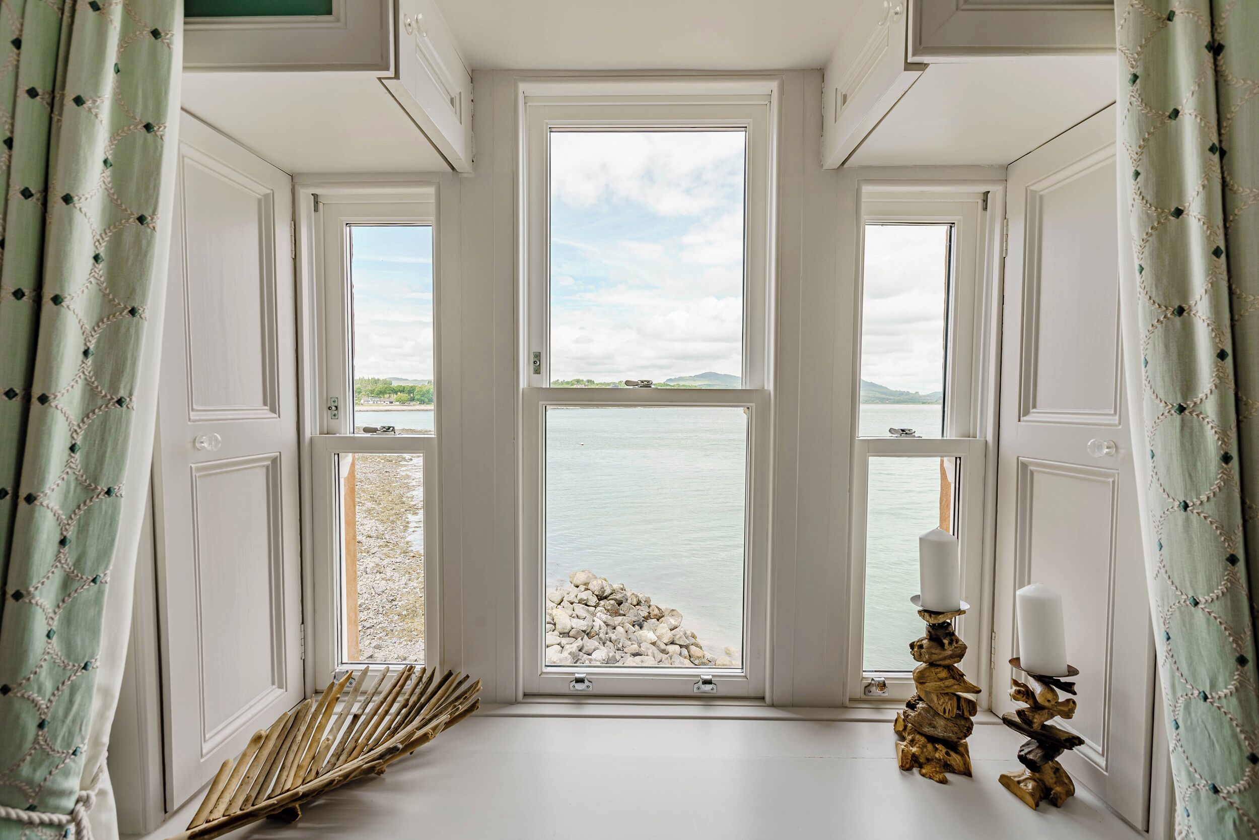 A room with a large window overlooking a rocky shoreline and a body of water. There are two candles on wooden stands on the windowsill.