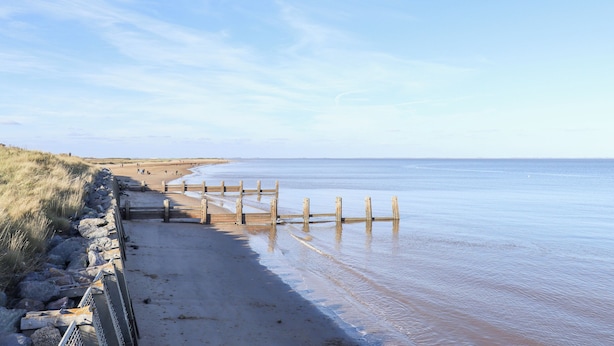 Une plage avec une jetée en bois qui s’avance dans des eaux calmes, un rivage sablonneux et des dunes herbeuses.