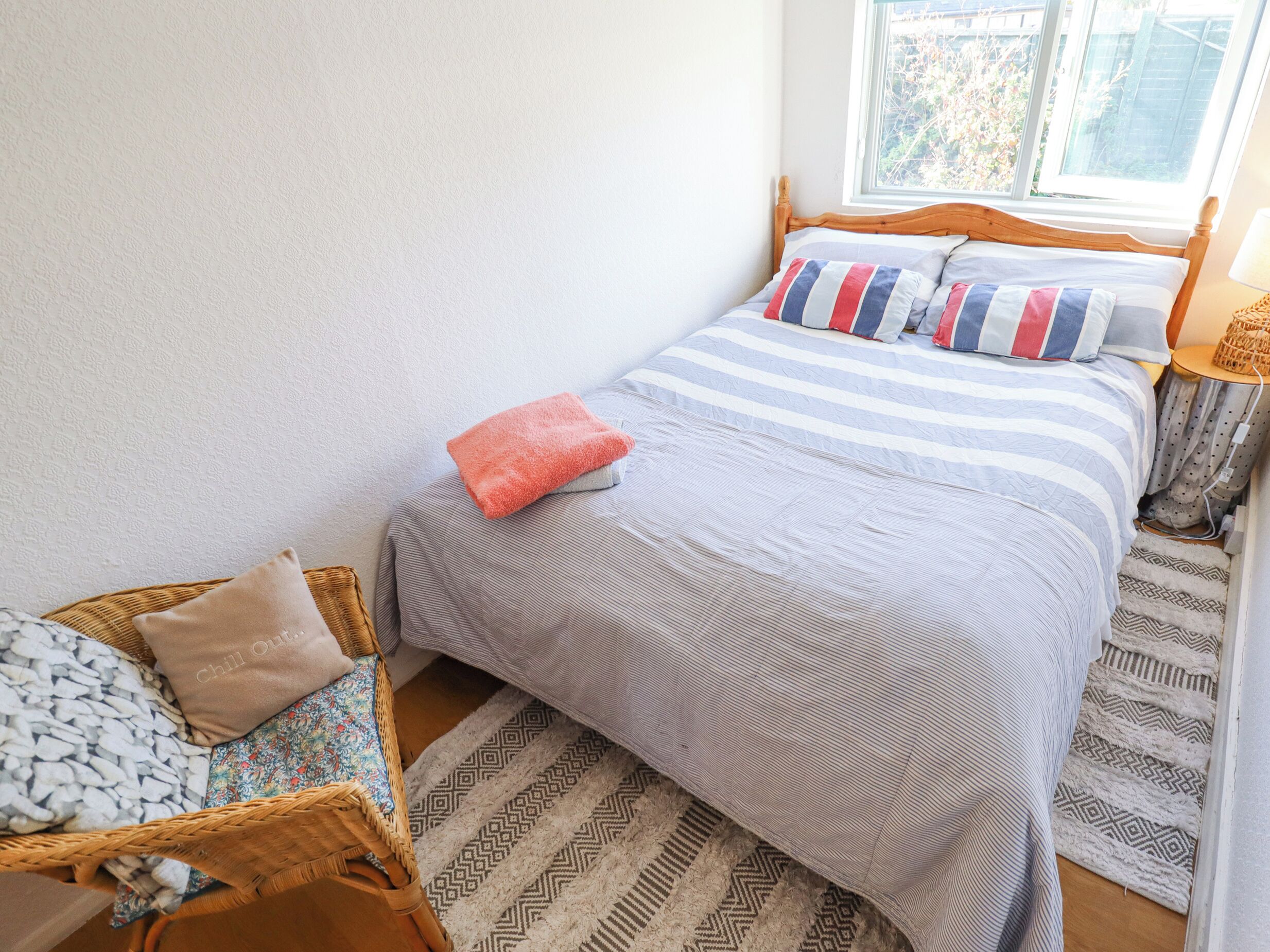 Un dormitorio ordenado con cama, una silla de mimbre y un tapete estampado.