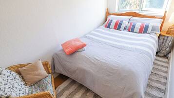 Un dormitorio ordenado con cama, una silla de mimbre y un tapete estampado.