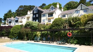 A swimming pool with a wooden deck and lounge chairs, surrounded by lush greenery and multi-story buildings. - 17 St Elmo Court (Salcombe)