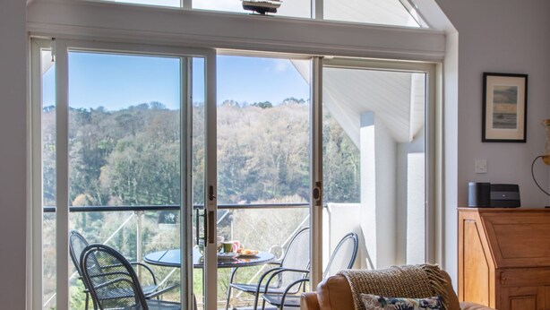 A room with a sliding glass door leading to a balcony with a table and chairs, a leather sofa, and a wooden cabinet. - 17 St Elmo Court (Salcombe)