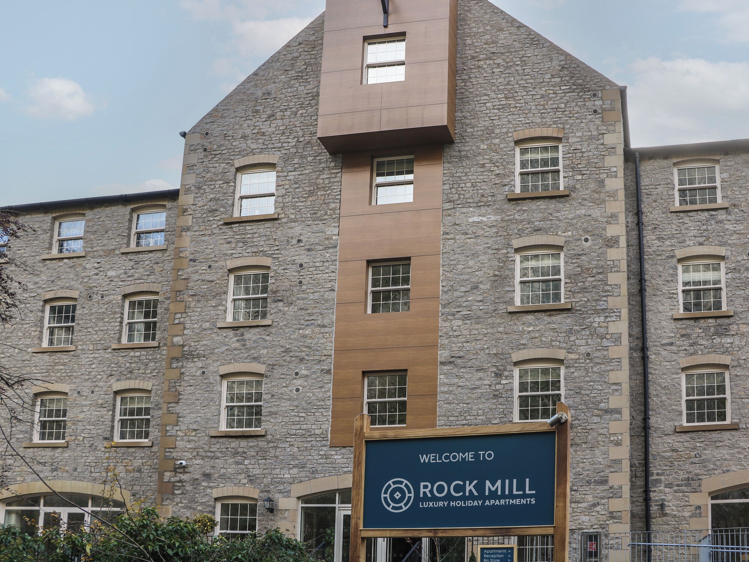 A multi-story building with a stone facade and a modern brown section. A sign reads "Welcome to Rock Mill Luxury Holiday Apartments."