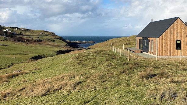 A wooden cabin with a sloped roof, situated on a grassy hill overlooking a coastal landscape. - Skye Falls Chalet (Glendale)