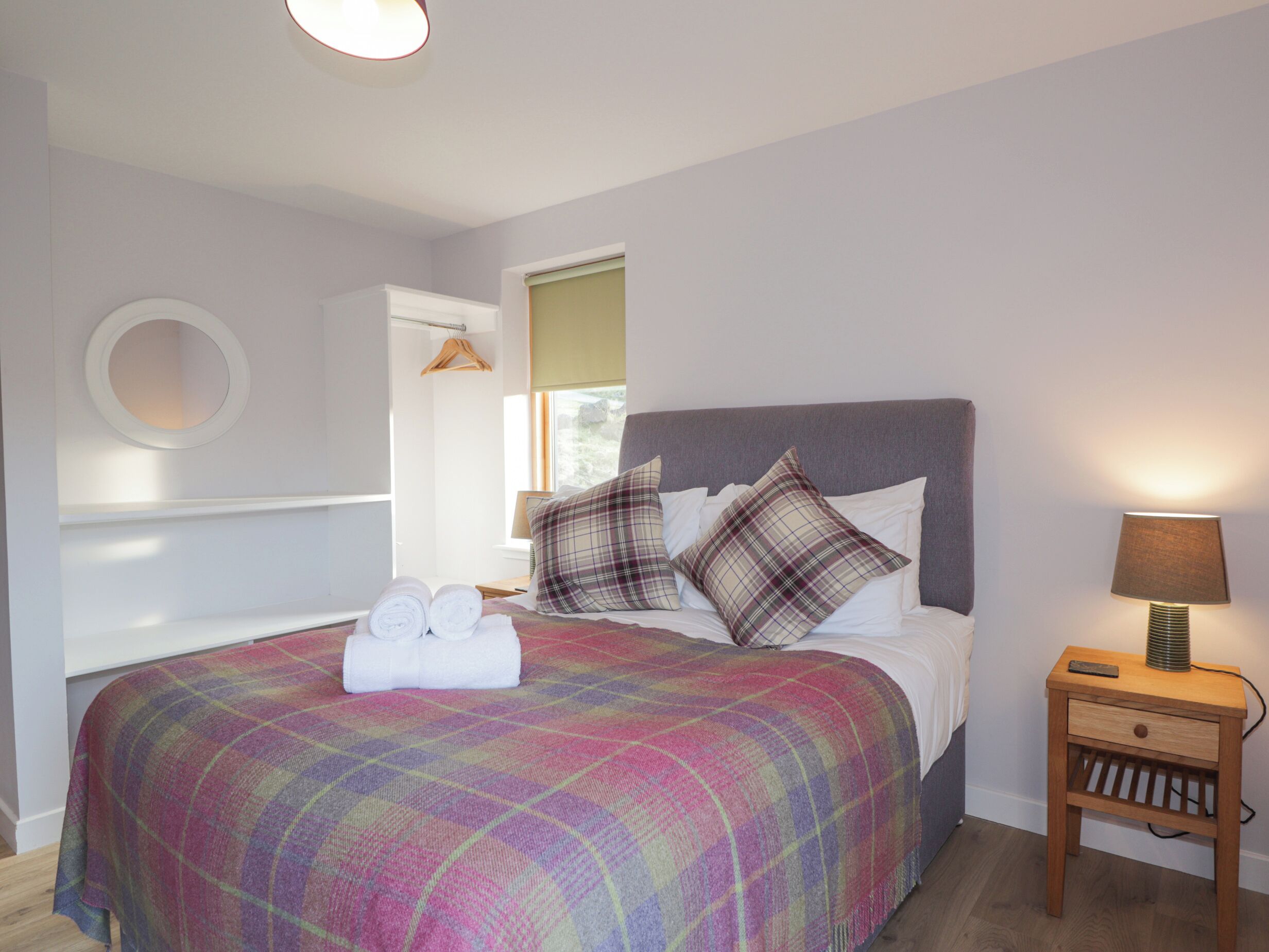 A bedroom with a bed, pillows, a bedside table, a lamp, a window, and a hanging garment rack.