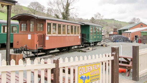 Ein historischer Zugwagen steht an einem Bahnhof, mit einem hölzernen Zaun und einem Schild für Golden Film Motoröl.
