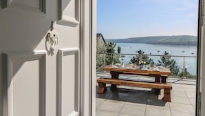 A balcony with a wooden table set for dining, overlooking a body of water with boats and hills in the distance. - Fronwig (New Quay)