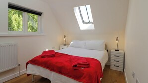 Ein Schlafzimmer mit Dachfenster, einem Bett mit roter Decke, Nachttischen mit Lampen und einem Fenster mit Blick auf Bäume.