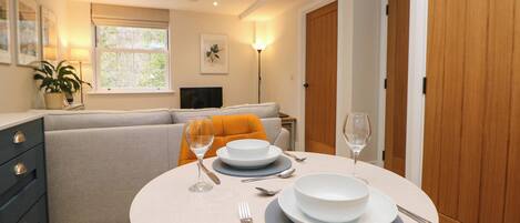 A dining area with a round table set for two, a sofa, and a TV in the background.