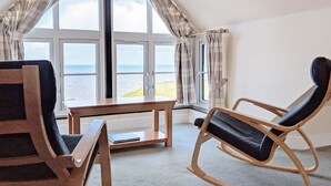 Ett rum med ett stort fönster med utsikt över havet, två gungstolar, ett litet bord och en vy över kustlinjen.