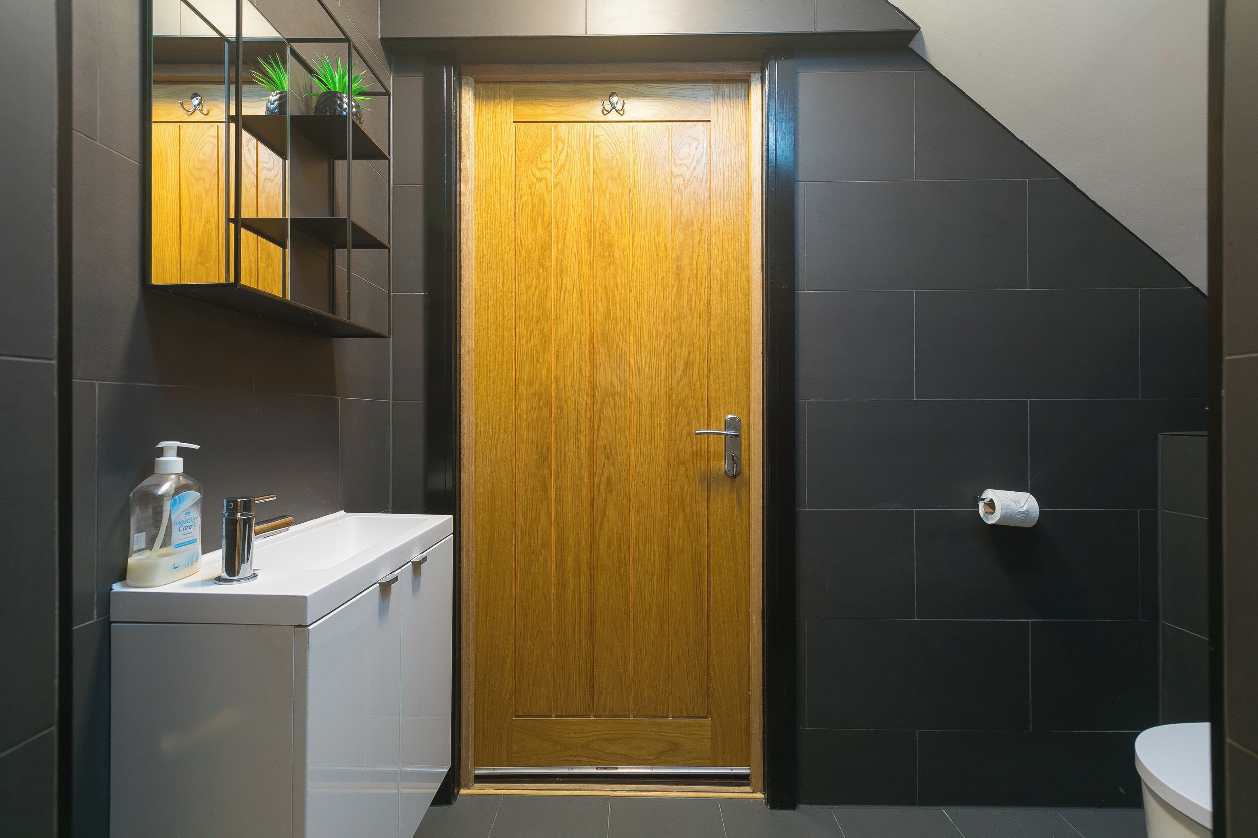 A bathroom with a wooden door, a white sink, a mirror, a shelf with plants, and a toilet.