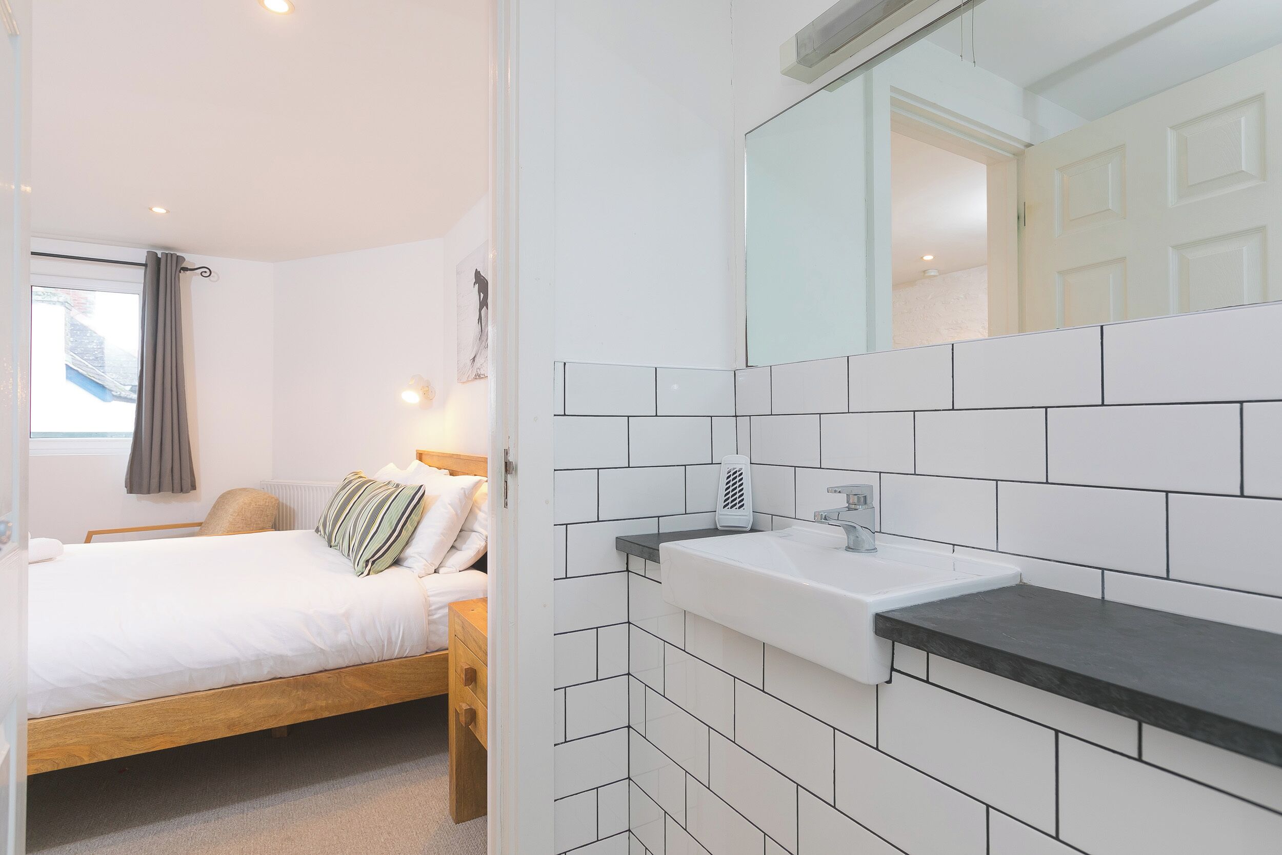 Ein Schlafzimmer mit einem Bett, einem Nachttisch und einem Schminktisch. Es gibt ein Badezimmer mit Waschbecken, Spiegel und Handtuchhalter.