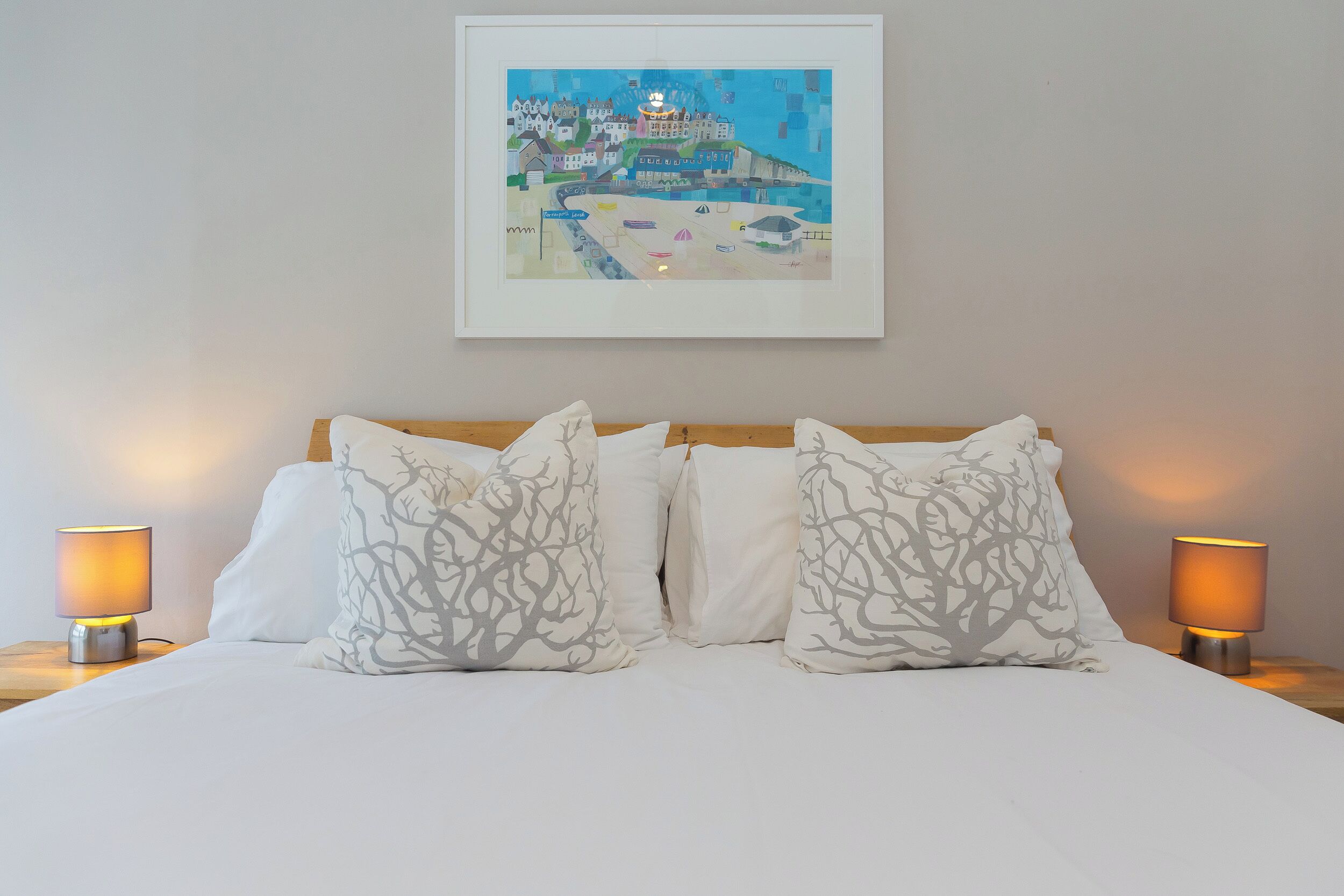 A neatly made bed with white bedding and patterned pillows, flanked by two bedside lamps. A framed artwork depicting a coastal scene hangs above the bed.