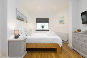 A bedroom with a wooden bed, white bedding, a bedside table with a lamp, a dresser, and two framed pictures on the wall.