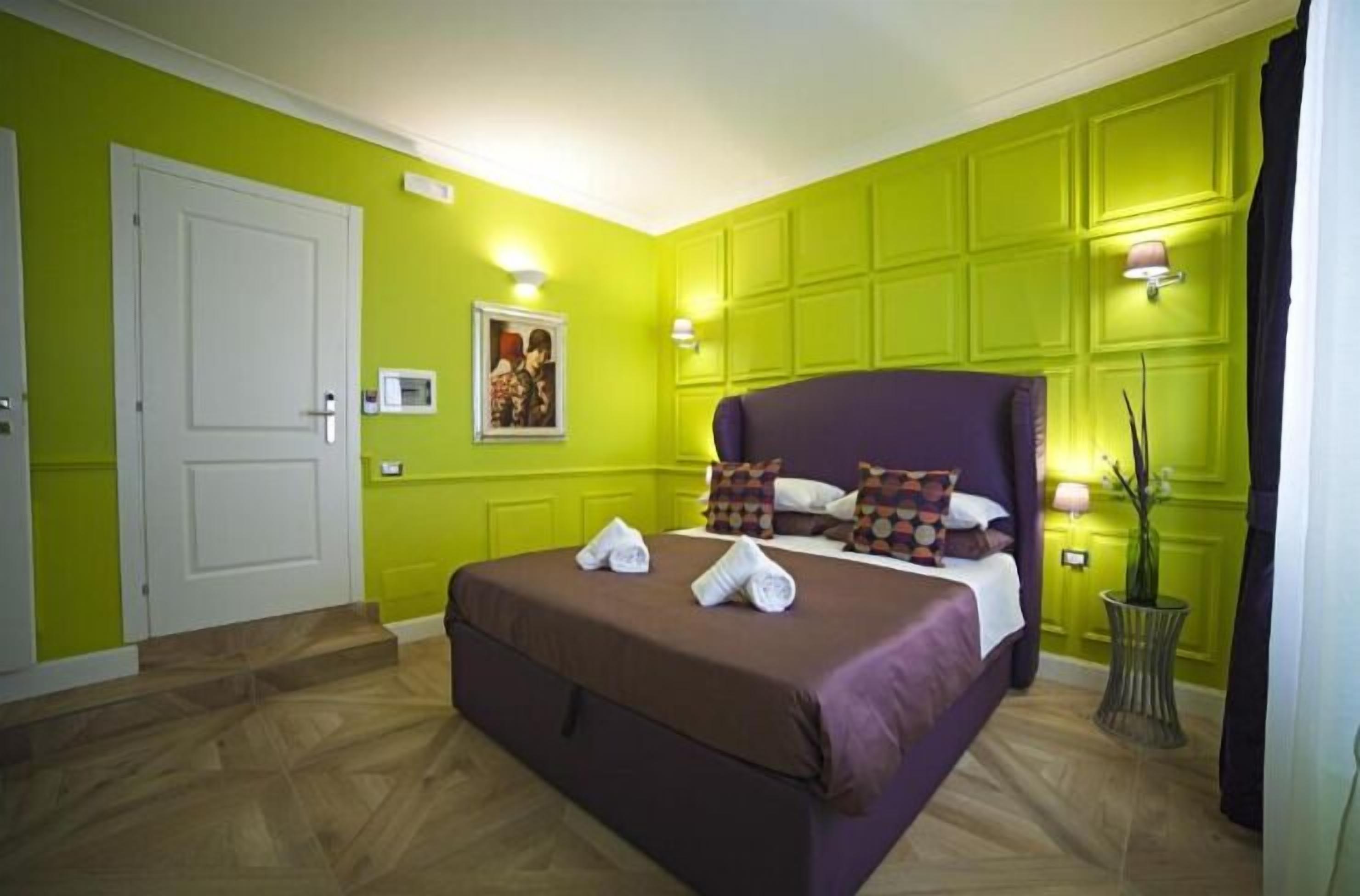 A room with a bed, a door, a wall-mounted picture, and a small table with a vase.