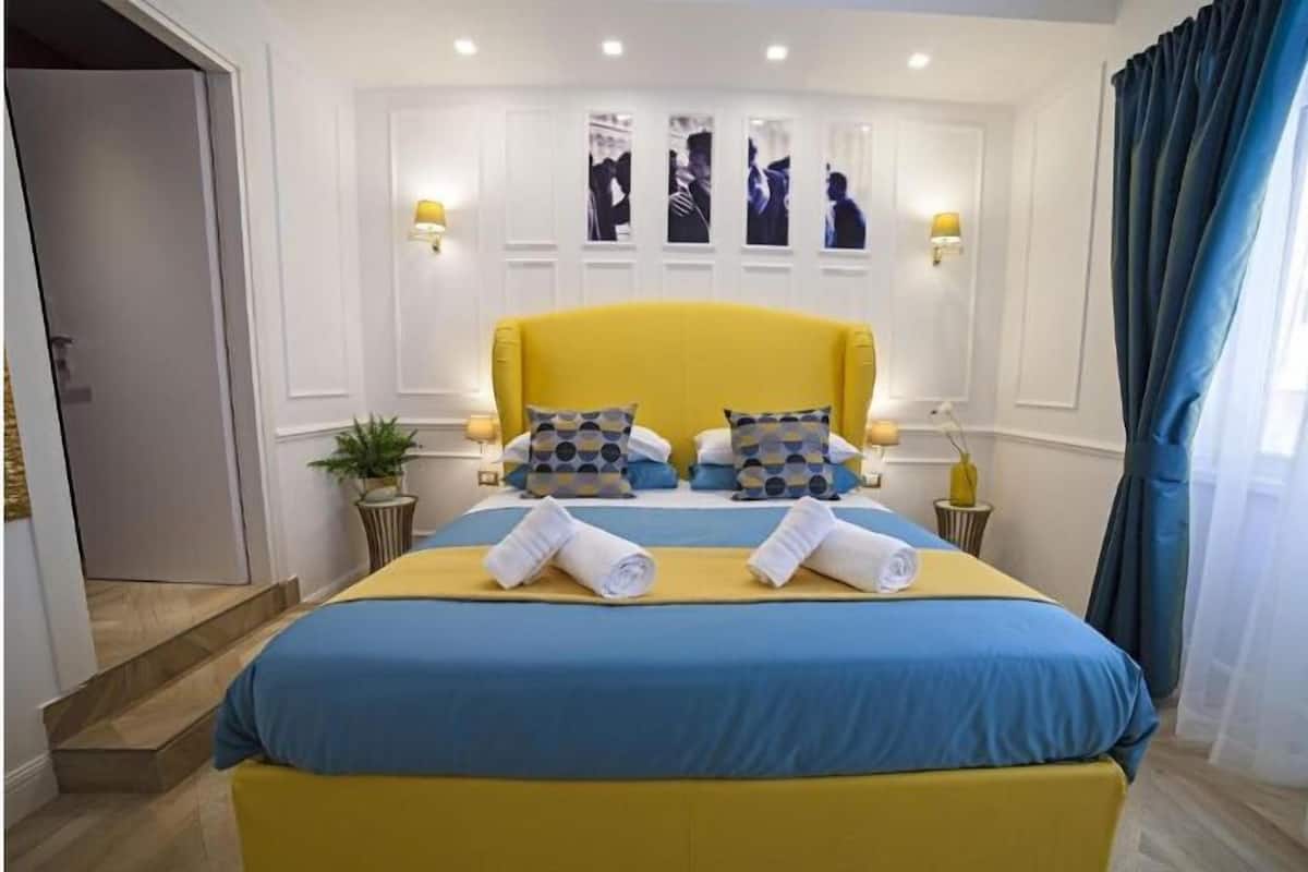 A hotel room with a large bed, two bedside lamps, a small table, and a potted plant.