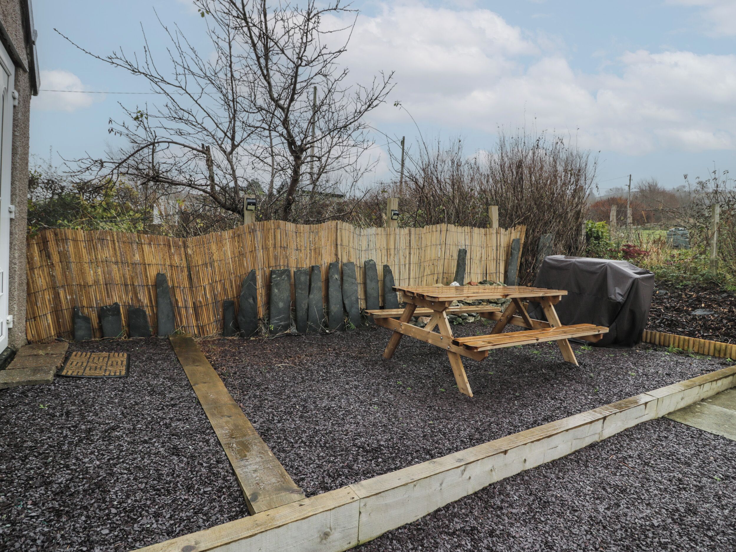 A wooden table and bench in a garden area with a bamboo fence and a covered grill.