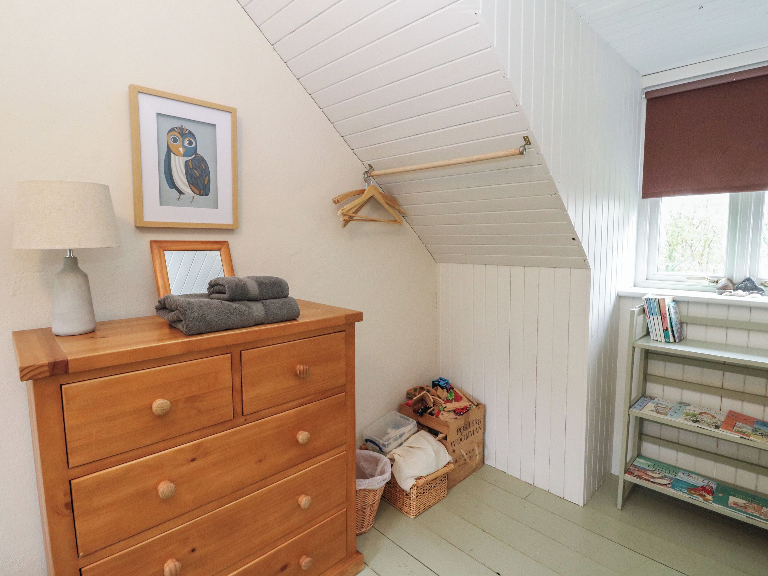 A cozy room with a wooden dresser, a framed owl picture, a lamp, and a window with books on a shelf.