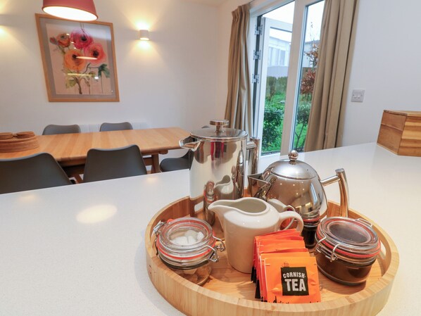 A breakfast setup with a tray of tea bags, a teapot, a jug, and a sugar bowl on a wooden table in a room with a dining area and a window with curtains. - Una, Lodge 73 (St Ives)