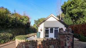 A small stone-walled building with a white door and windows, surrounded by greenery and a paved path. - Studio Apartment (Minehead)