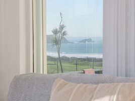 A room with a view of the ocean through a window, a bed, and a fence in the background.