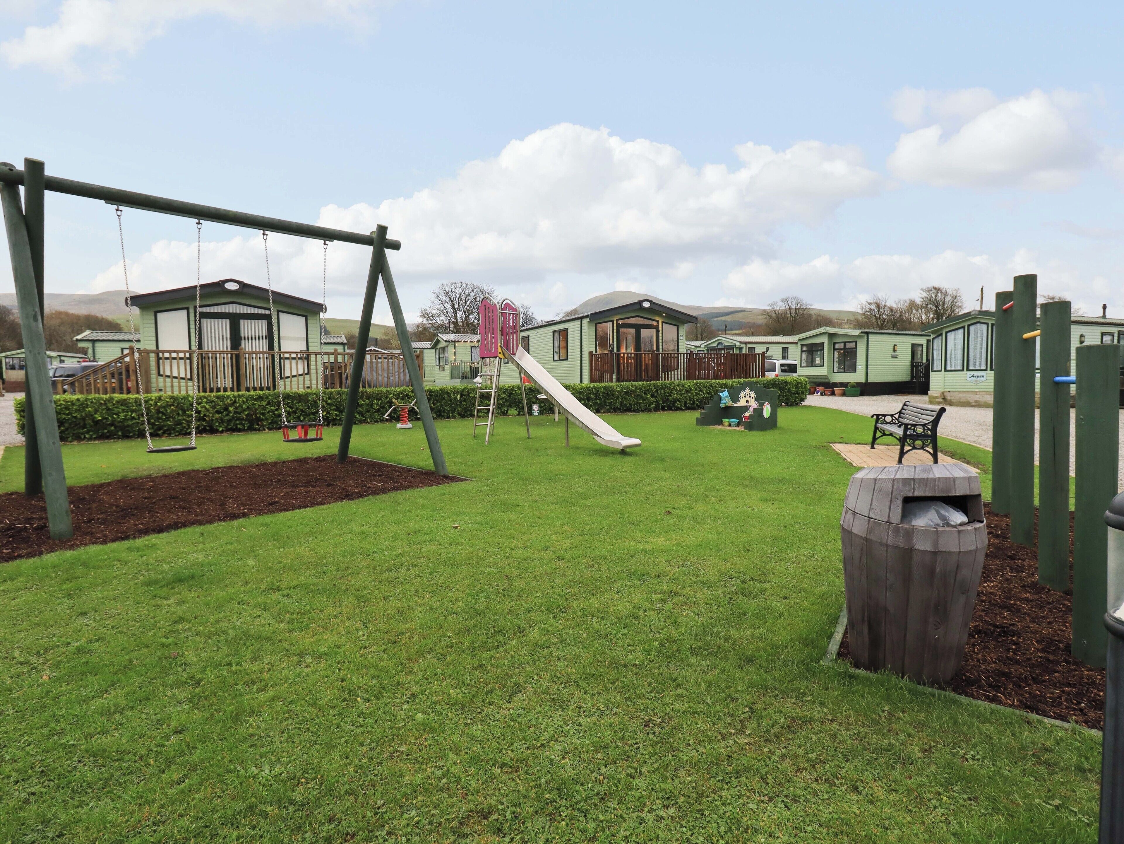 A playground with swings and a slide, surrounded by green grass and a wooden fence.
