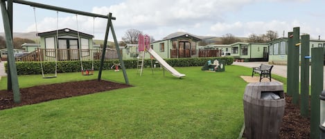 A playground with swings and a slide, surrounded by green grass and a wooden fence.
