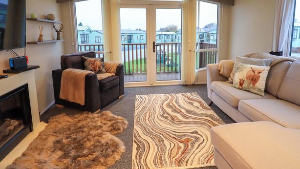 A living room with a fireplace, a sofa, a large window with a view of houses, and a television on a shelf. - Highlands Way Lodge (Workington)