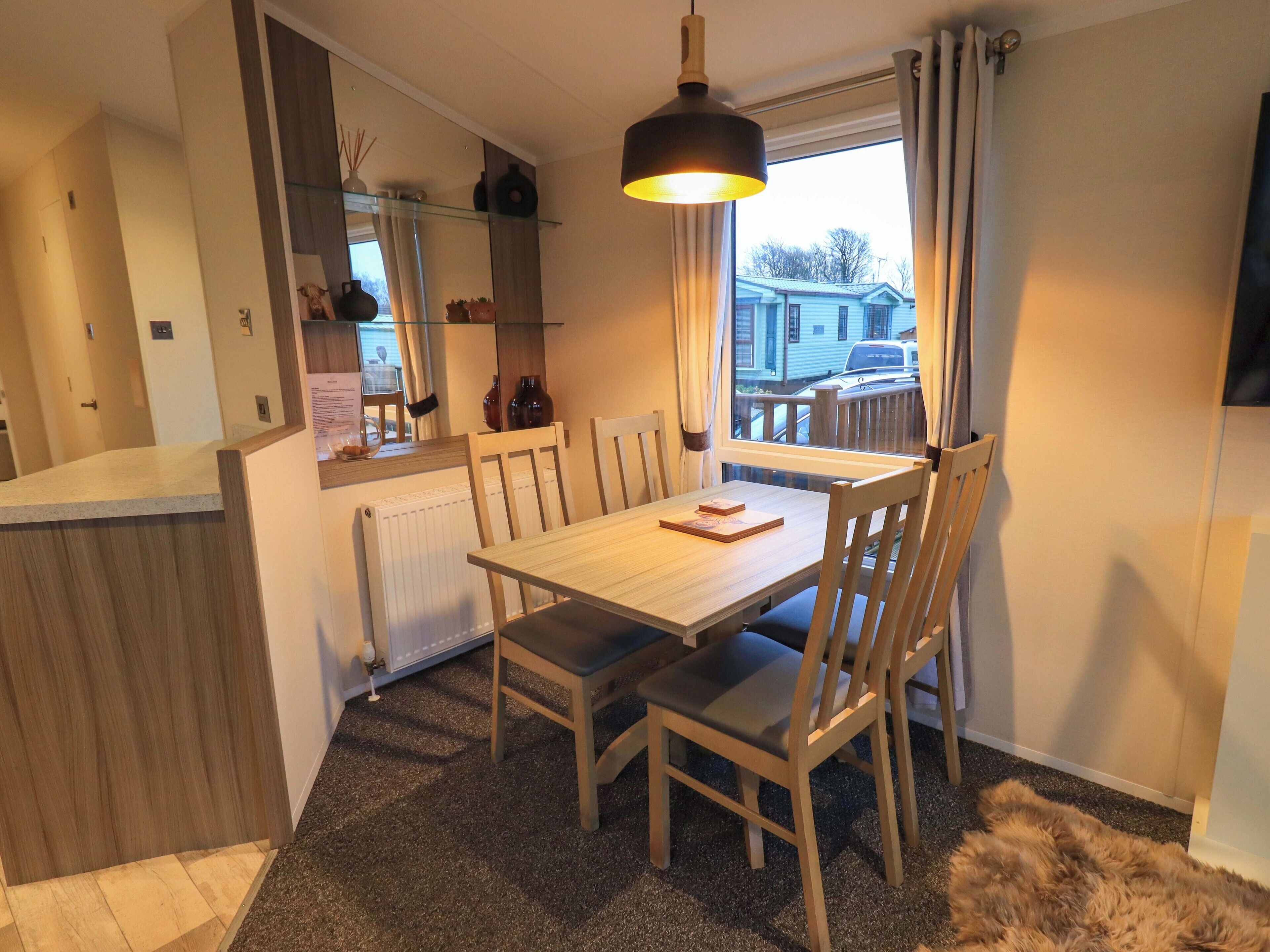 A dining area with a wooden table and chairs, a hanging light fixture, a window with curtains, and a view of a residential street.