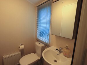 A small bathroom with a toilet, a sink, a mirror, and a window with blinds. - Highlands Way Lodge (Workington)