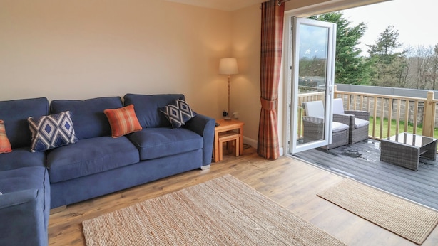 Un salón con un sofá azul, cojines estampados, una mesita de madera con una lámpara y un gran ventanal corredera que da a una terraza exterior con mobiliario de mimbre.