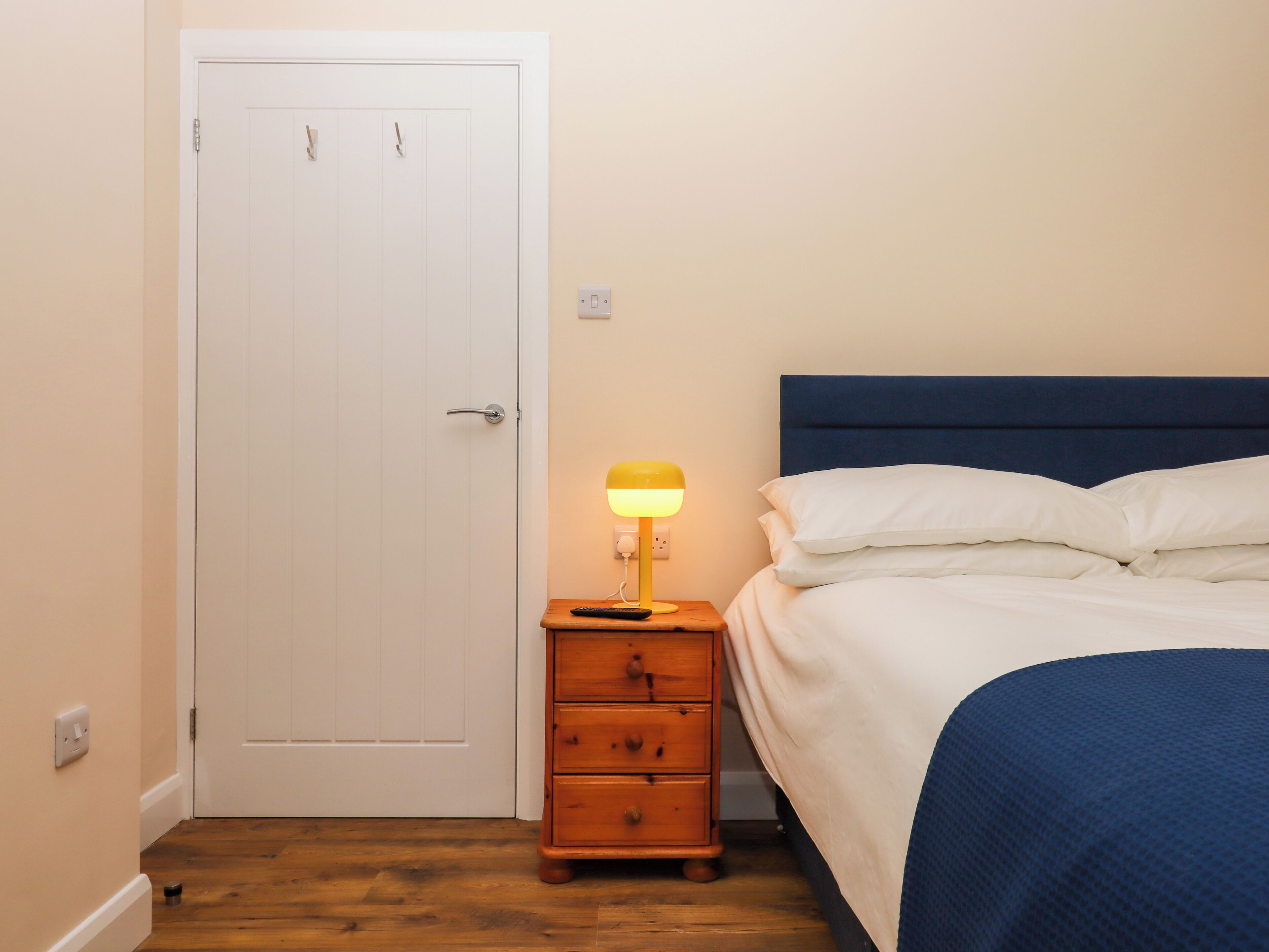 A bedroom with a white door, a bedside table with a lamp, a wooden dresser, and a bed with a blue headboard and white bedding.