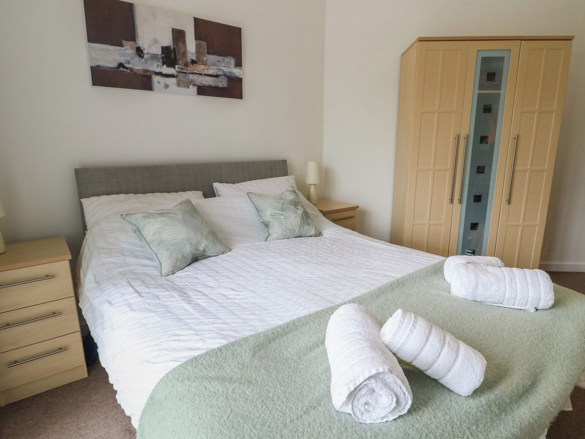 A neatly made bed with pillows, a green throw blanket, and rolled towels on a green carpet. There is a wooden wardrobe with glass doors and a small bedside table with a lamp.