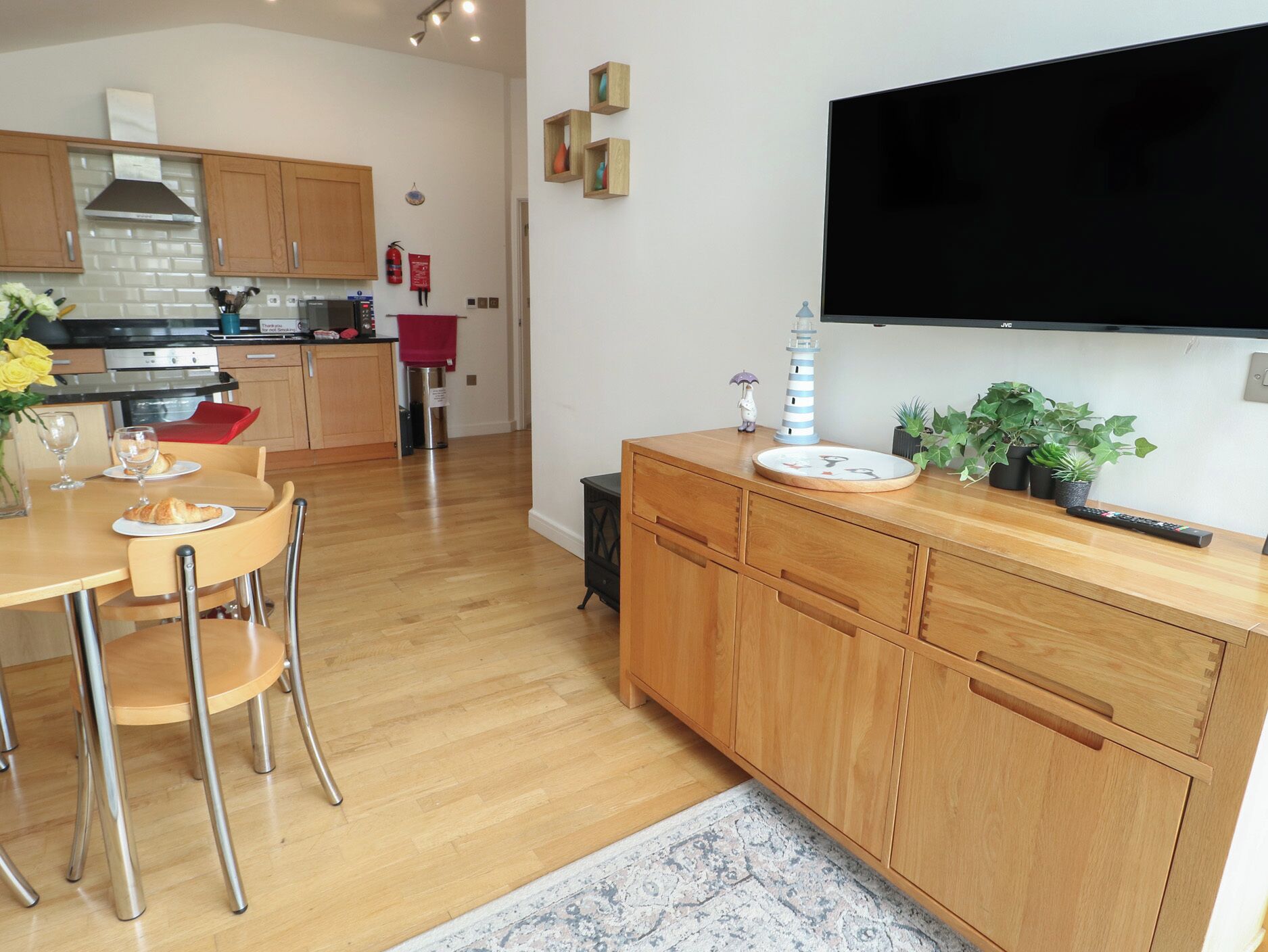 A modern kitchen with wooden cabinets, a dining table with chairs, a TV mounted on the wall, and a small potted plant on the counter.