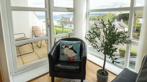 A corner room with a black leather chair, a potted plant, and a view of the sea and houses through large windows. - Seaview (Tyn-y-Gongl)