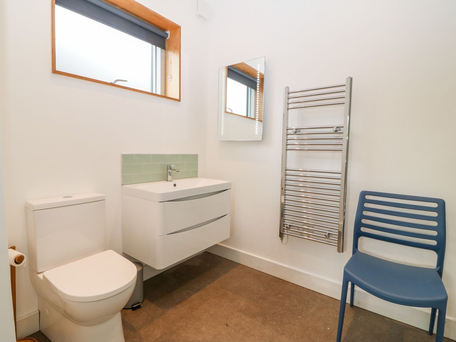 Un baño con inodoro, lavamanos, espejo, toallero y una silla.