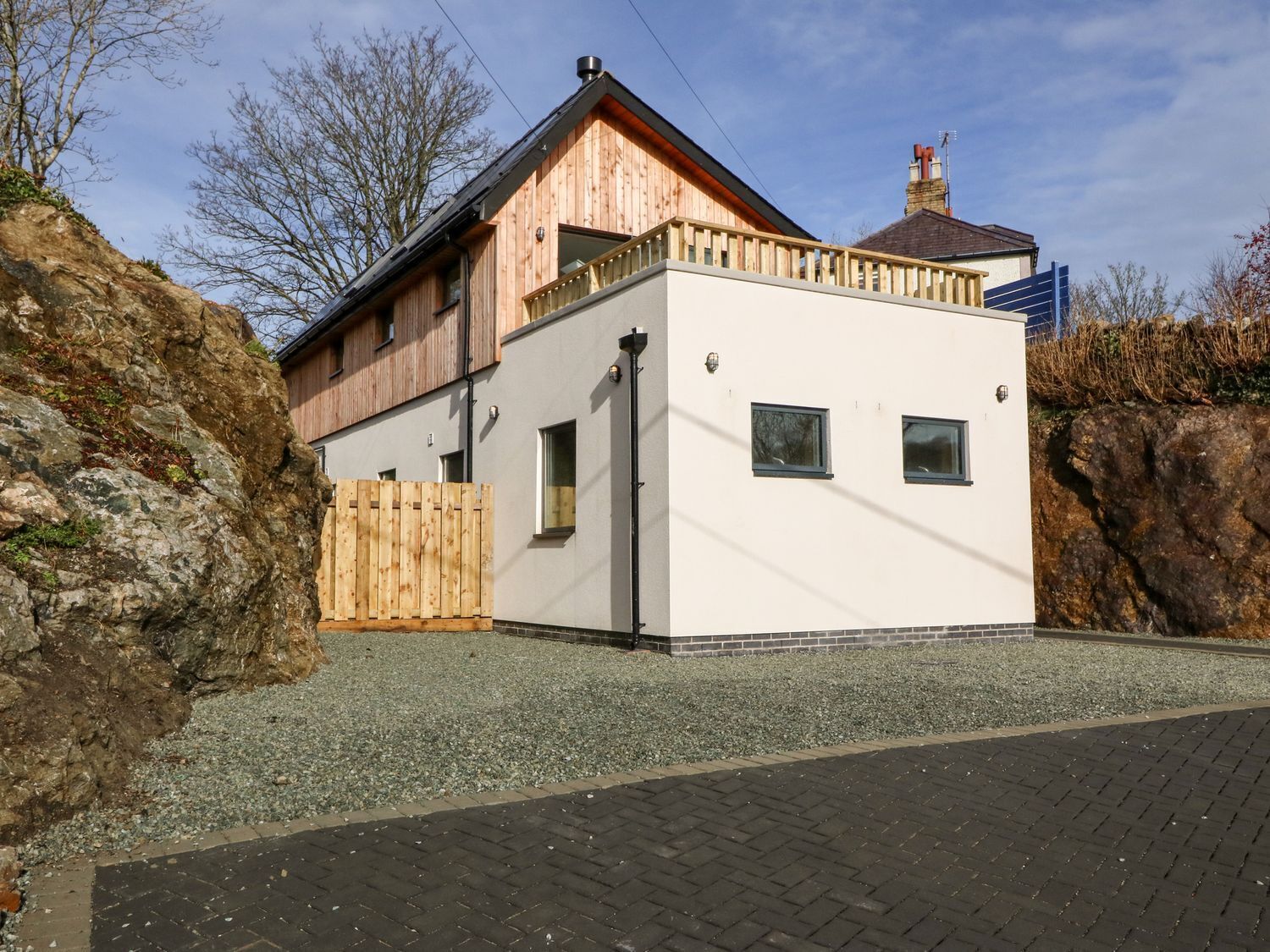 A modern two-story house with a wooden extension, a white facade, and a gravel driveway.