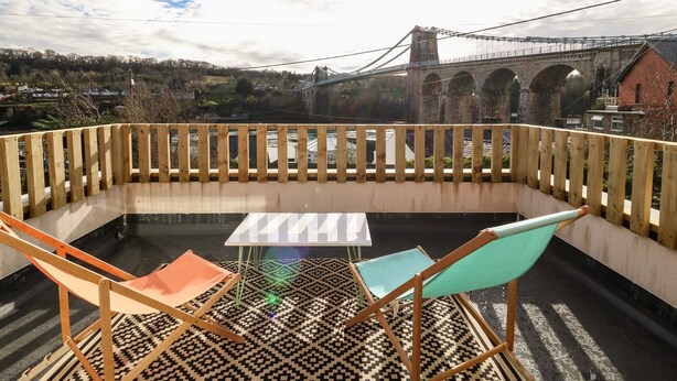 A rooftop terrace with a table and two chairs, overlooking a historic bridge and a town. - Ty Chwarel (Bangor)
