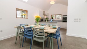 A dining area with a table set for six, surrounded by blue chairs. A vase with yellow flowers is on the table. The room has a high ceiling with skylights and a kitchen in the background. - Ty Chwarel (Bangor)