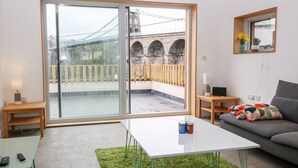 A modern living room with a large sliding glass door leading to a balcony, a grey sofa, a white coffee table, and a small wooden side table. - Ty Chwarel (Bangor)