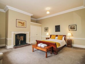 A bedroom with a fireplace, a bed with yellow and white bedding, two bedside tables with lamps, and framed pictures on the walls. - Caynham Court (Ludlow)