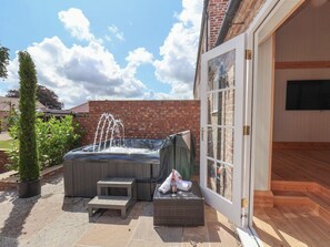 Un jacuzzi en un patio trasero con un muro de ladrillo, una terraza de madera y un ventanal que da a una habitación con televisor.