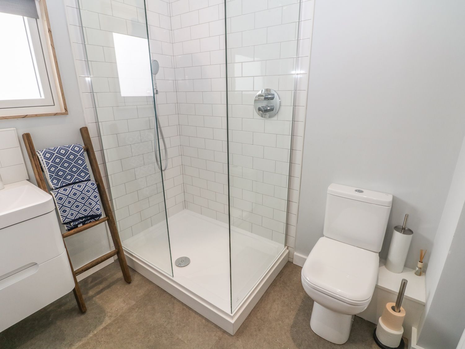 A modern bathroom with a glass-enclosed shower, white tiles, a toilet, and a wooden chair with a patterned cushion.