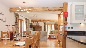 A dining area with wooden chairs and a table set for four, a living room with a sofa and a coffee table, and a kitchen with a fire blanket and a clock. - Swallows Reach (Totnes)