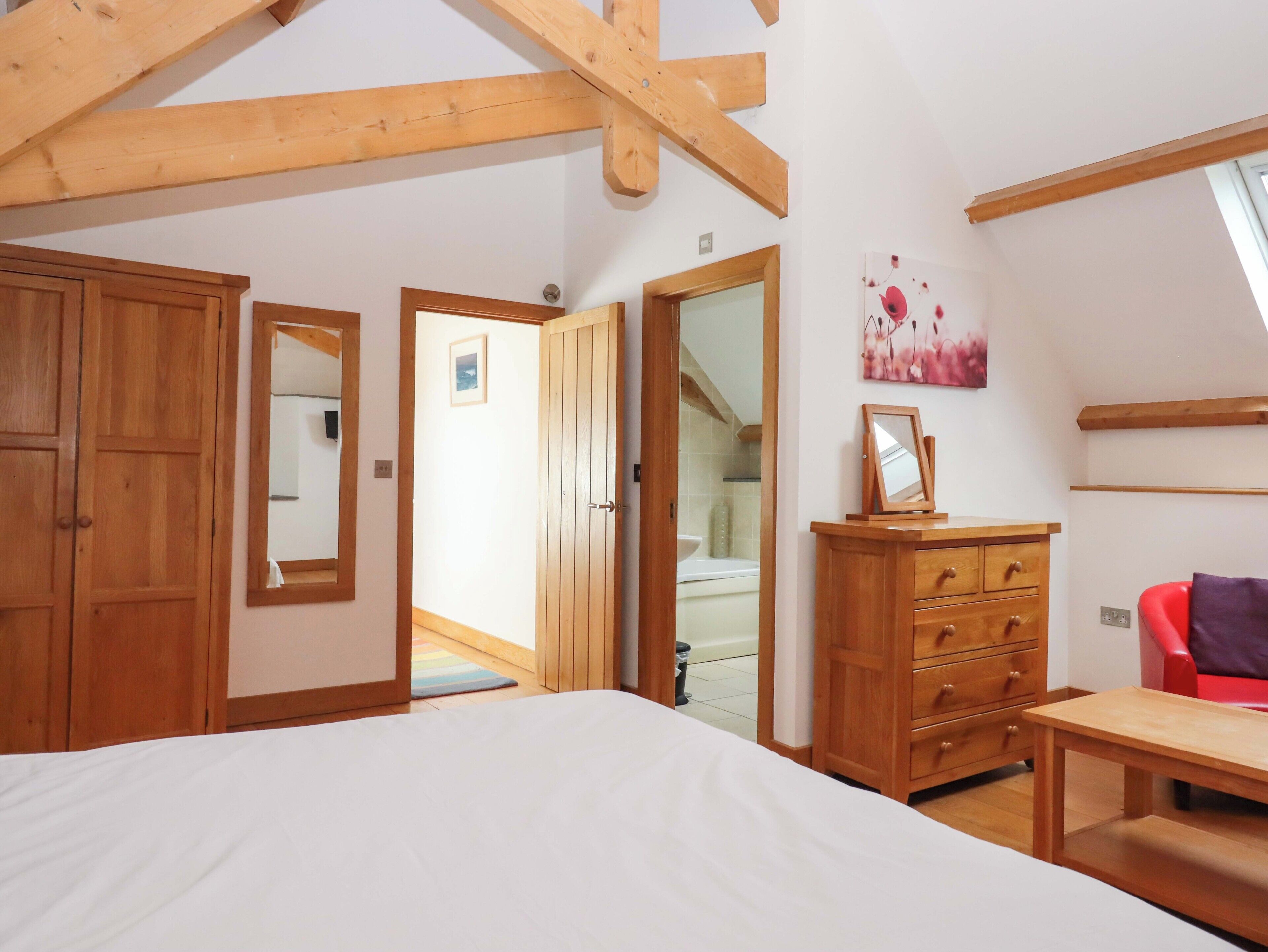 A bedroom with wooden beams, a bed, a dresser, a mirror, and a red chair.