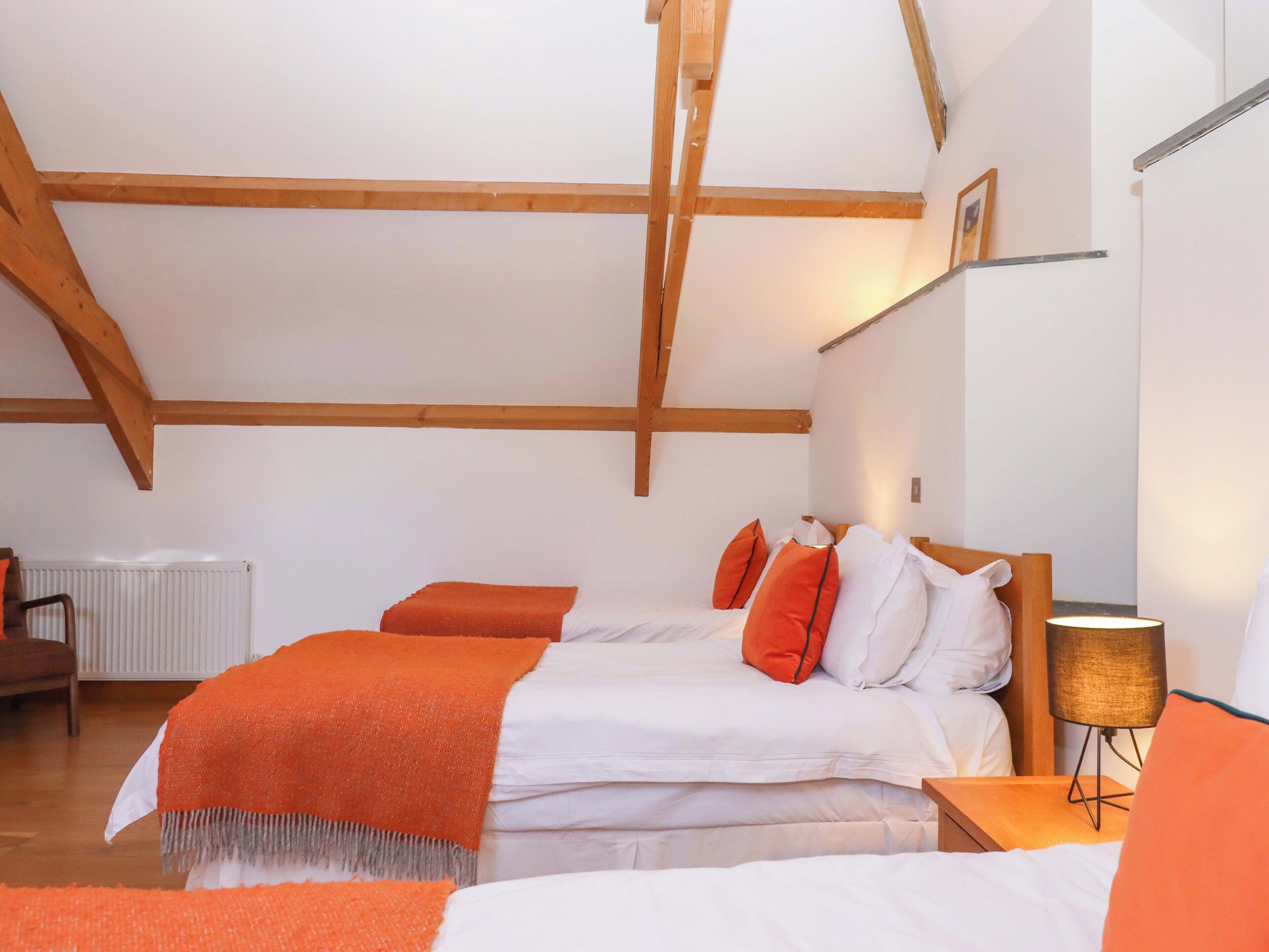Una camera da letto con soffitto spiovente, travi in legno, un letto con biancheria arancione e bianca, una lampada da comodino e un termosifone.