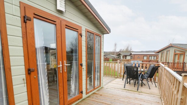 A wooden deck with a table and chairs, a sliding glass door, and a view of neighboring houses. - 6 Sherwood (Carnforth)