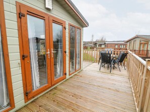 A wooden deck with a table and chairs, a sliding glass door, and a view of neighboring houses. - 6 Sherwood (Carnforth)