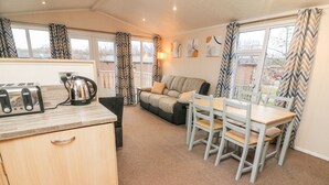A compact living space with a kitchenette, a sofa, a dining table, and a view through large windows. - 6 Sherwood (Carnforth)