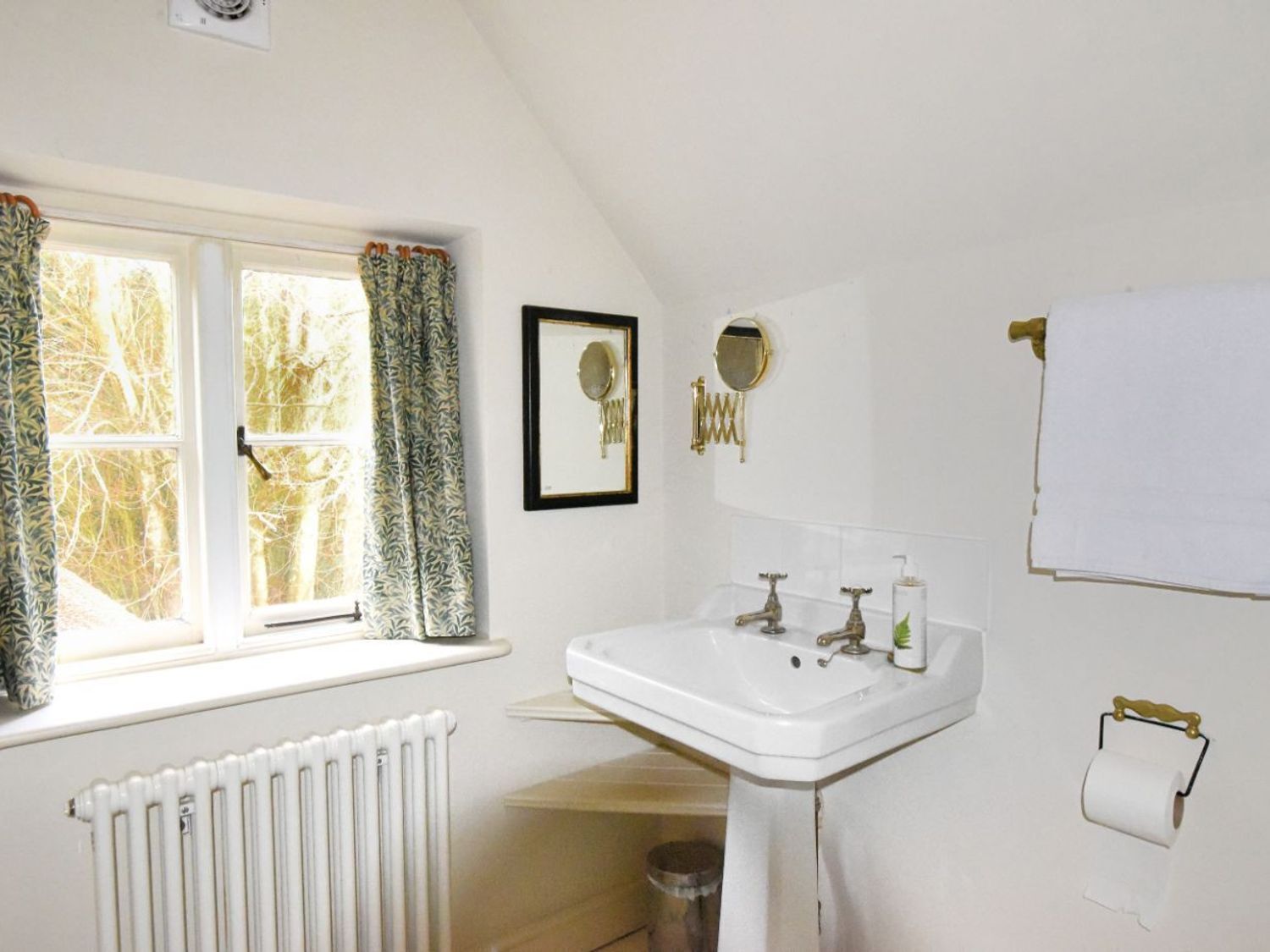 Une salle de bain avec un lavabo blanc, deux miroirs, un radiateur et une fenêtre avec des rideaux.